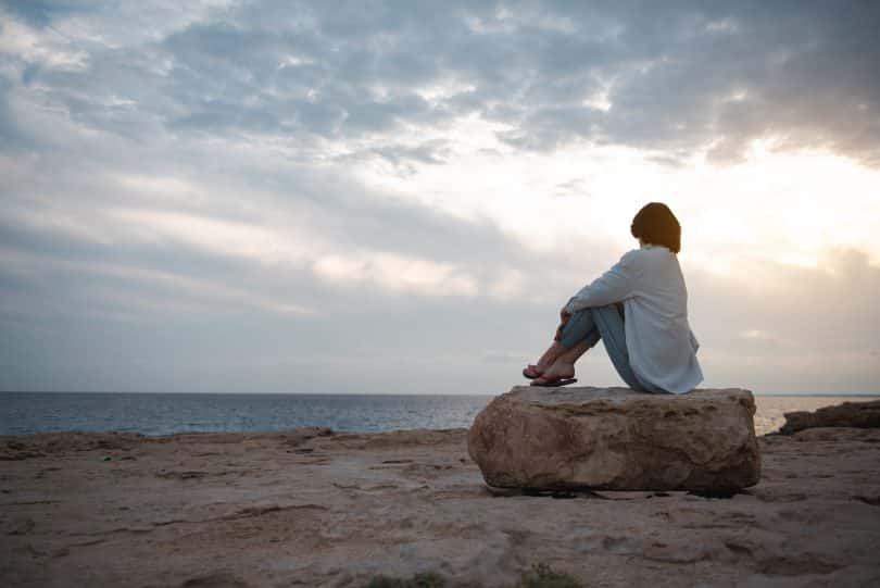 Mulher pensativa sentada em uma pedra da praia olhando para o mar