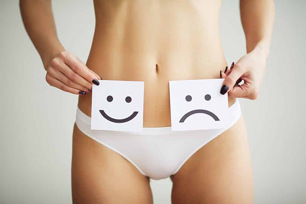 Imagem do corpo de uma mulher - do seio para baixo. Ela usa uma calcinha branca e segura sobre a sua barriga duas placas de emotions: um alegre e outro em dúvida.