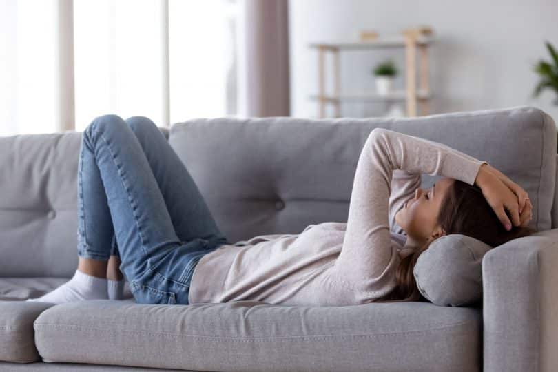 Mulher deitada em sofá com as pernas flexionadas e as mãos na cabeça. Ela parece irritada e exausta.