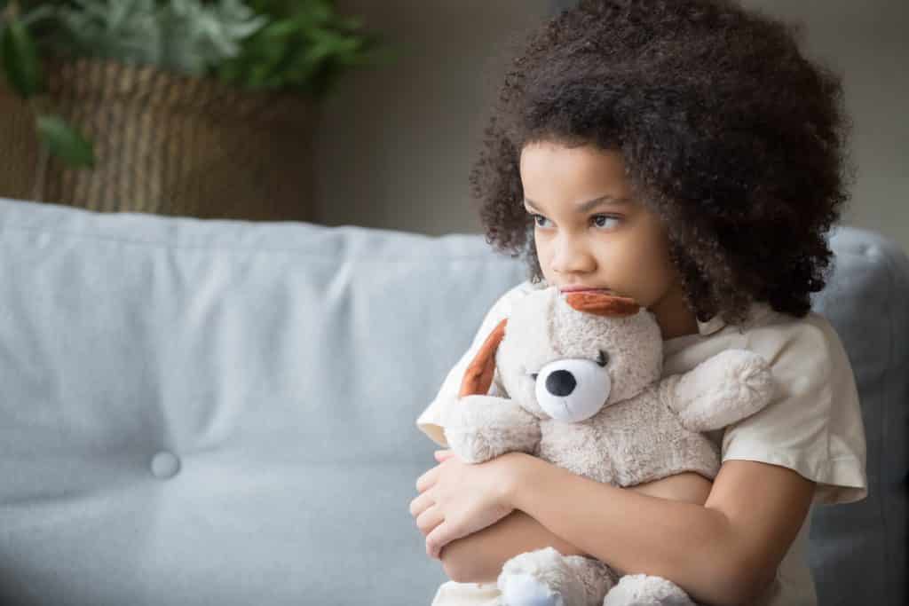 Imagem de uma criança afrodescendente sentada em um sofá de couro branco. Ela segura em seu colo um ursinho de pelúcia bege.