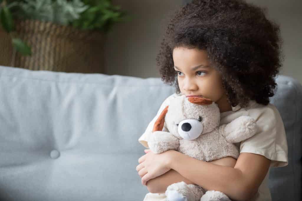 Imagem de uma criança afrodescendente sentada em um sofá de couro branco. Ela segura em seu colo um ursinho de pelúcia bege.
