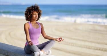 Mulher de cabelos cacheados sentada na beira da praia, meditando com os olhos fechados.