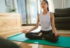 Mulher sentada, com as pernas cruzadas, meditando no chão de uma sala.