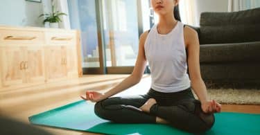 Mulher sentada, com as pernas cruzadas, meditando no chão de uma sala.