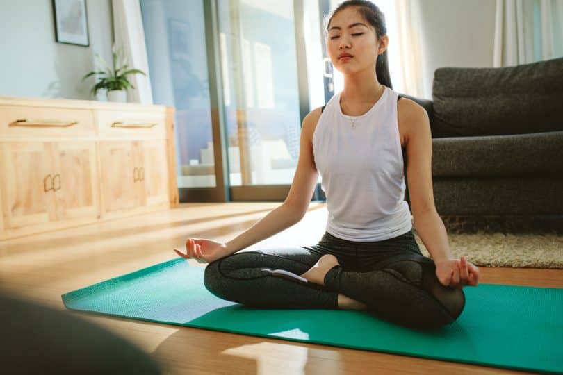 Mulher sentada, com as pernas cruzadas, meditando no chão de uma sala.