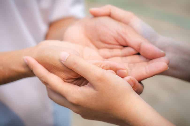 As mãos de duas pessoas. Todas com as palmas viradas para cima, enquanto uma das pessoas segura as mãos da outra por baixo.