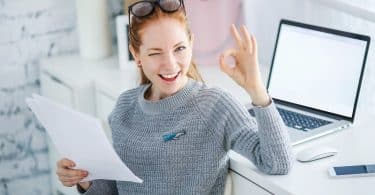 Mulher trabalhando em mesa de Home Office com notebook e celular enquanto segura papeis de escritório e sorri.