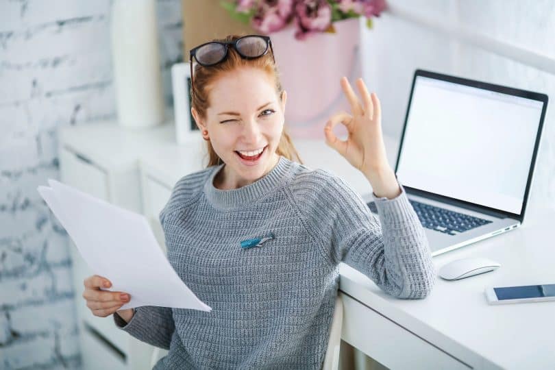 Mulher trabalhando em mesa de Home Office com notebook e celular enquanto segura papeis de escritório e sorri.