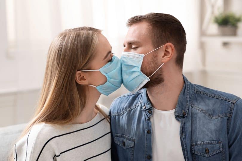 Casal em quarentena, beijando-se com máscaras faciais de proteção.