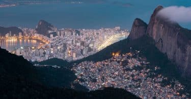 Vista aérea do Rio de Janeiro, do lado direito, uma favela acesa em topo de morro, enquanto na parte de baixo, perto do mar, prédios grandes de classe alta.