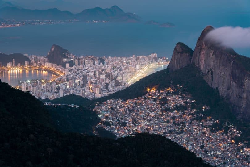 Vista aérea do Rio de Janeiro, do lado direito, uma favela acesa em topo de morro, enquanto na parte de baixo, perto do mar, prédios grandes de classe alta.