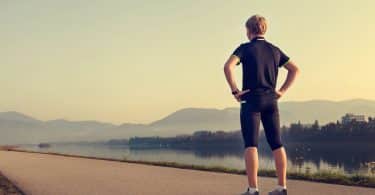 Homem com roupas de corrida parado em pé, de costas, no meio de uma estrada.