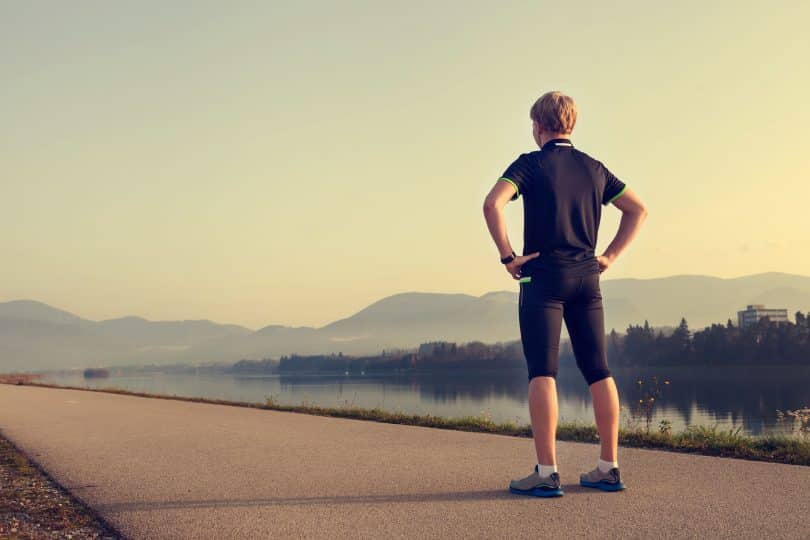 Homem com roupas de corrida parado em pé, de costas, no meio de uma estrada.