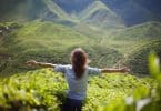 Mulher de costas sentada em montanha com área verde ao fundo e braços abertos