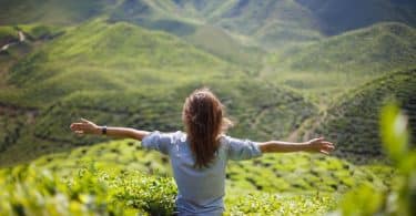 Mulher de costas sentada em montanha com área verde ao fundo e braços abertos