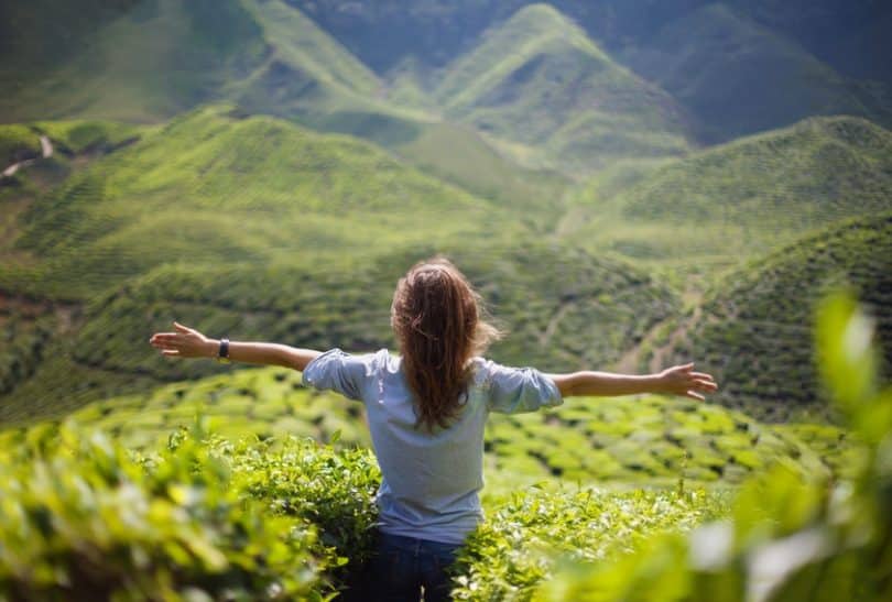 Mulher de costas sentada em montanha com área verde ao fundo e braços abertos