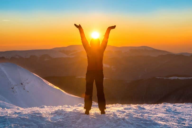 Pessoa com os braços erguidos em meio a neve. Ela está no pico de uma montanha.