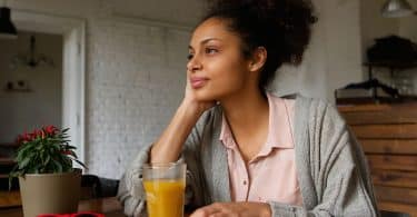 Mulher sentada na mesa da cozinha segurando um suco e olhando para frente pensativa