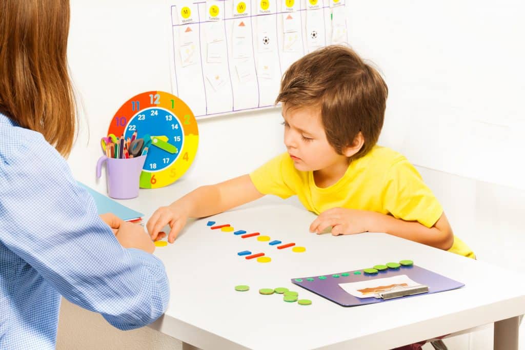 Imagem de uma criança jogando e montando um quebra-cabeça. Ela está sentada de frente com uma mulher que está observando-a. A mesa é branca e sobre ela tem os jogos infantis e um relógio de parede colorido.