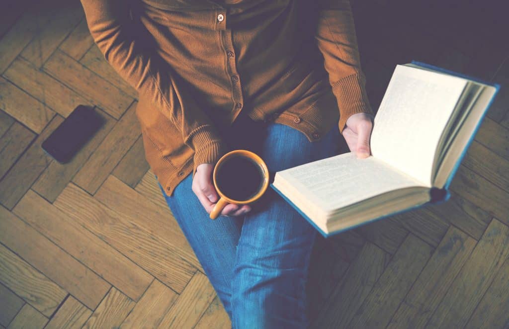 Mulher sentada no chão segurando um livro aberto com uma mão, e uma xícara de café com a outra mão.