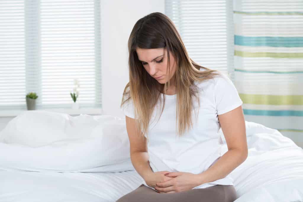 Mulher dentro do quarto, sentada sobre a sua cama. Ela está com dores no útero e suas mãos estão sobre ele. Ela usa uma camiseta branca e  uma calça leggin.
