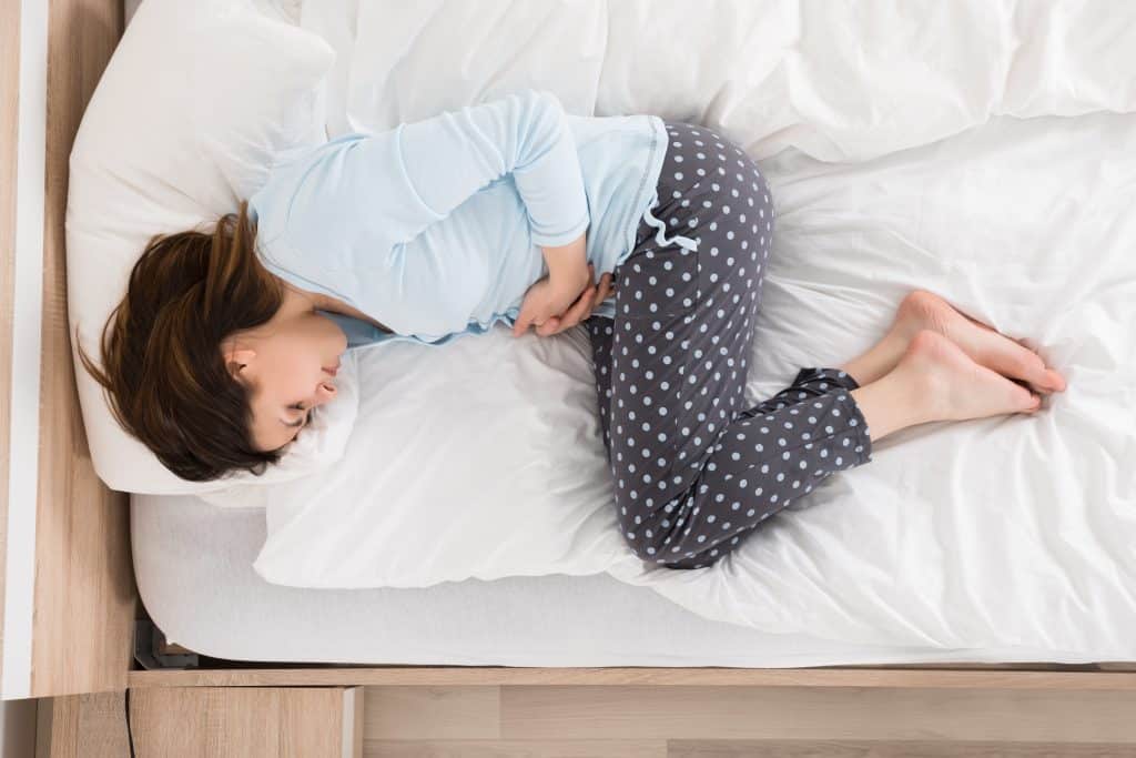 Mulher deitada sobre a sua cama. Ela veste um pijama de frio cinza e azul claro. Ela está com dores próximo ao útero e suas mãos estão sobre ele.