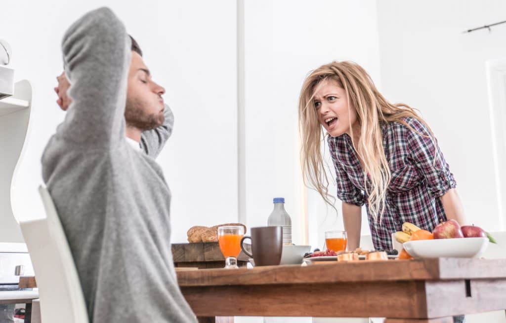 Mulher gritando com homem, que coloca as mãos atrás da cabeça, na mesa de café da manhã.