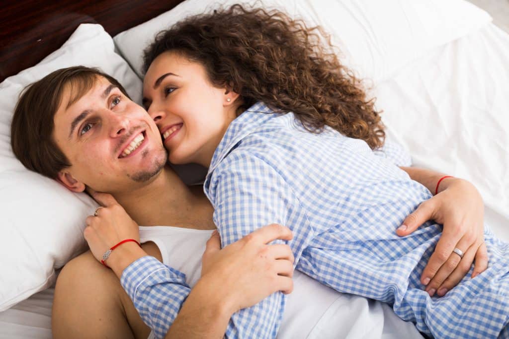 Homem e mulher deitados em cama, abraçados e sorrindo.