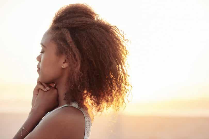 Mulher de cabelos cacheados vista de perfil, com os olhos fechados e as mãos junto ao queixo.