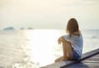 Garota sentada em ponte olhando para o lago com sol ao fundo
