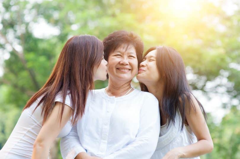 Duas mulheres beijando a mãe na bochecha, enquanto esta sorri.