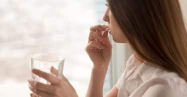 Mulher vista de perfil olhando por uma janela enquanto toma uma pílula de remédio junto a um copo d'água.