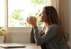 Mulher segurando uma xícara de chá e olhando para janela sentada em sua mesa da cozinha