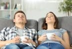 Homem e mulher, sentados em um sofá, cada um segurando uma caneca, relaxados no encosto, com os olhos fechados.