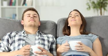 Homem e mulher, sentados em um sofá, cada um segurando uma caneca, relaxados no encosto, com os olhos fechados.