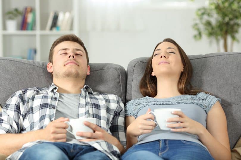 Homem e mulher, sentados em um sofá, cada um segurando uma caneca, relaxados no encosto, com os olhos fechados.