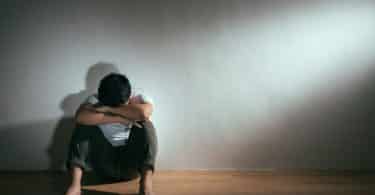 Homem sentado no chão, encostado em uma parede, com os joelhos flexionados e os braços apoiados neles. Ele esconde seu rosto apoiando a testa nos braços.