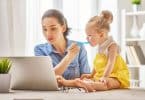 Mãe trabalhando com um computador em home office enquanto dá comida na boca de sua filha pequena, que senta em cima da mesa.