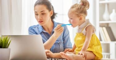 Mãe trabalhando com um computador em home office enquanto dá comida na boca de sua filha pequena, que senta em cima da mesa.