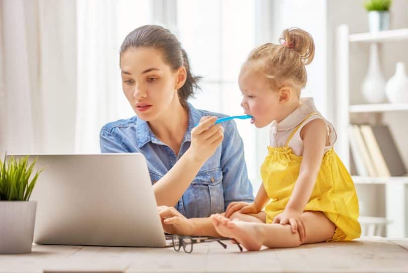 Mãe trabalhando com um computador em home office enquanto dá comida na boca de sua filha pequena, que senta em cima da mesa.