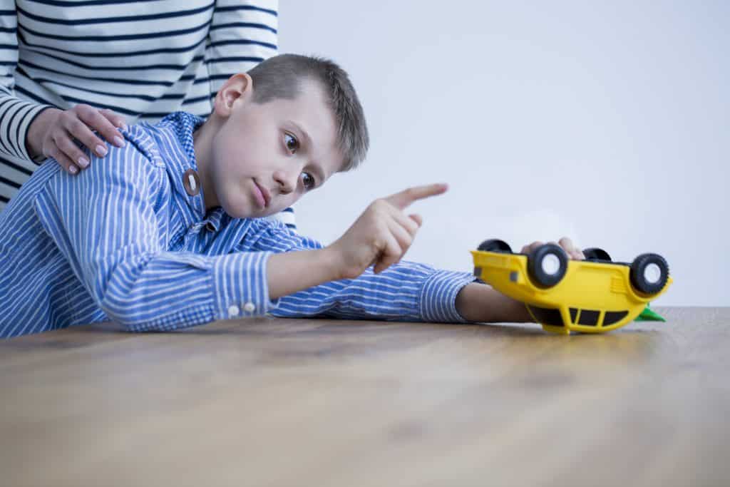 Imagem de um garoto olhando e apontando para o seu carrinho de brinquedo amarelo. Atrás dele, está a sua mãe colocando as mãos sobre os ombros.
