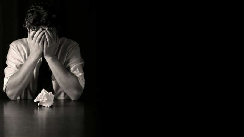 Imagem de um homem com as mãos no rosto em um cenário escuro, apoiado na mesa, como se estivesse tendo pensamentos ruins