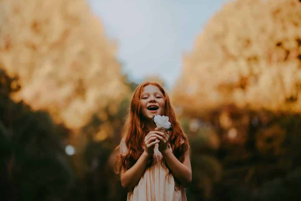 Menina em parque, segurando uma flor próxima ao seu rosto, e sorrindo com a boca aberta.