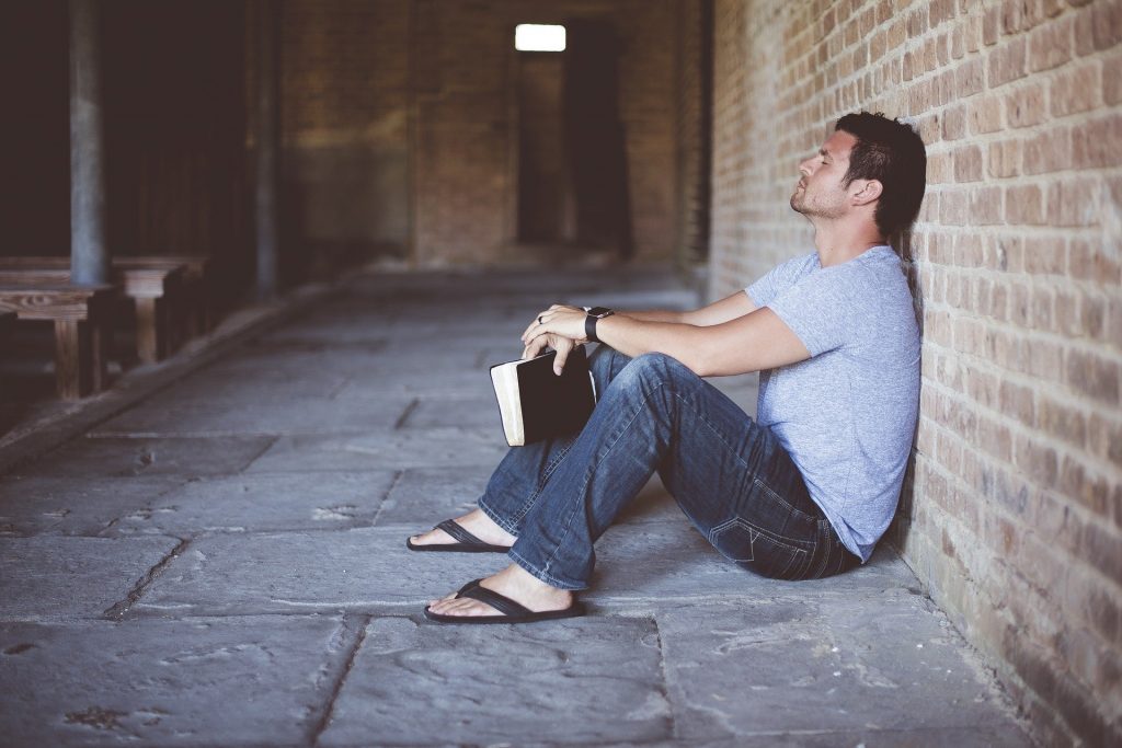 Imagem de um  homem sentado sozinho no chão, segurando entre as suas mãos um livro de capa preta. Ele está encostado em uma parede de tijolos e está pensativo.