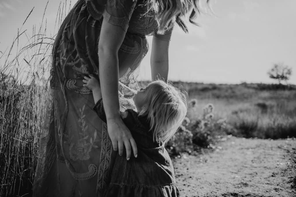 Mulher grávida de vestido pegando sua filha menor do colo, em um campo.