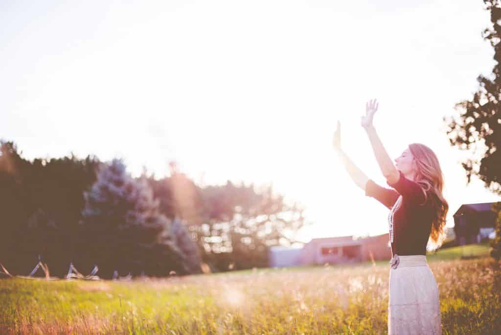 Mulher em campo gramado, com as mãos erguidas para o alto e para frente, em direção a luz do sol.