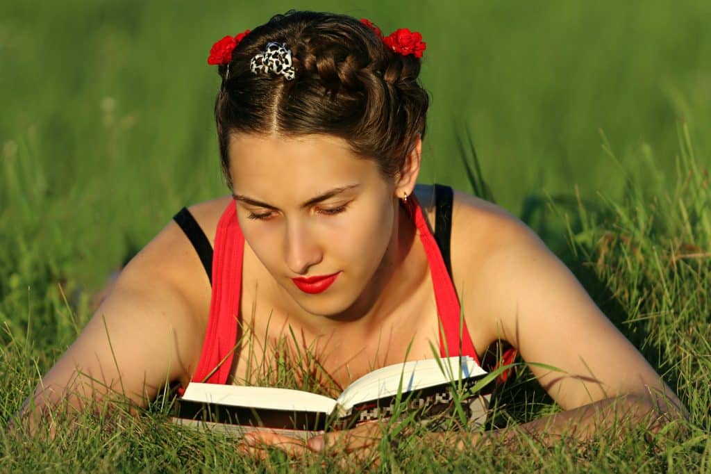 Imagem de uma garota em um parque. Ela está deitada sobre uma grama  e está lendo um livro.