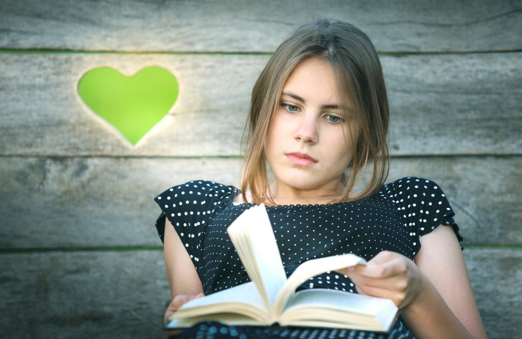 Menina sentada segurando entre as mãos um livro. Ela está pensando e folheando o livro. Ao fundo uma parede de madeira e ao lado a imagem de um coração pintado na cor verde.
