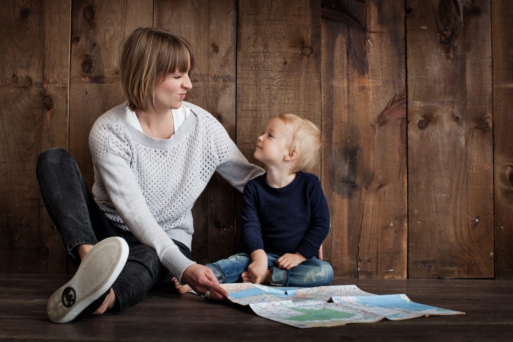 Mulher e menino bebê sentados lado a lado, em frente a um mapa. A mulher faz uma careta para o menino, que sorri.
