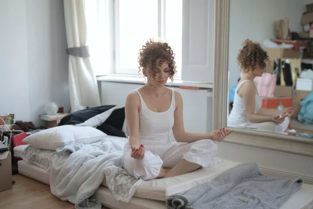 Mulher sentada em um quarto, de pernas cruzadas, meditando.